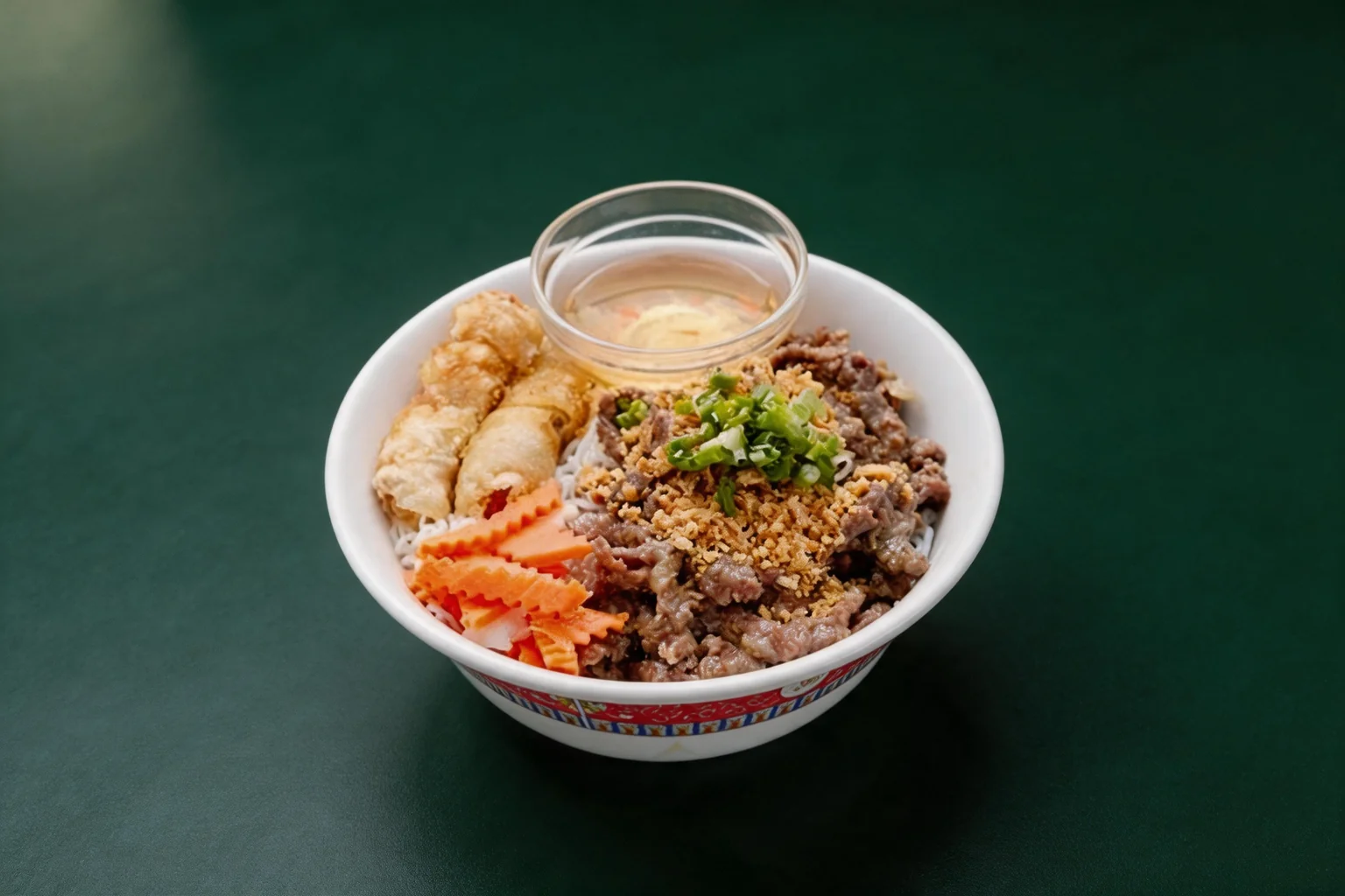 Bobun in a white bowl with a red border: vermicelli, beef, spring rolls, carrots and a dish of sauce, on a dark green background