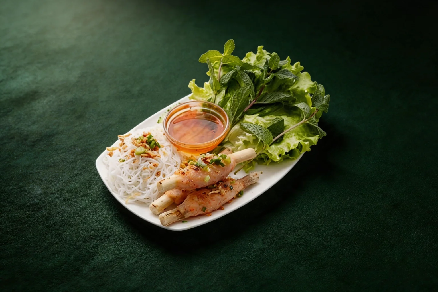 Sugar cane prawns on a white plate, vermicelli, lettuce, herbs and a dish of sauce, on a dark green background